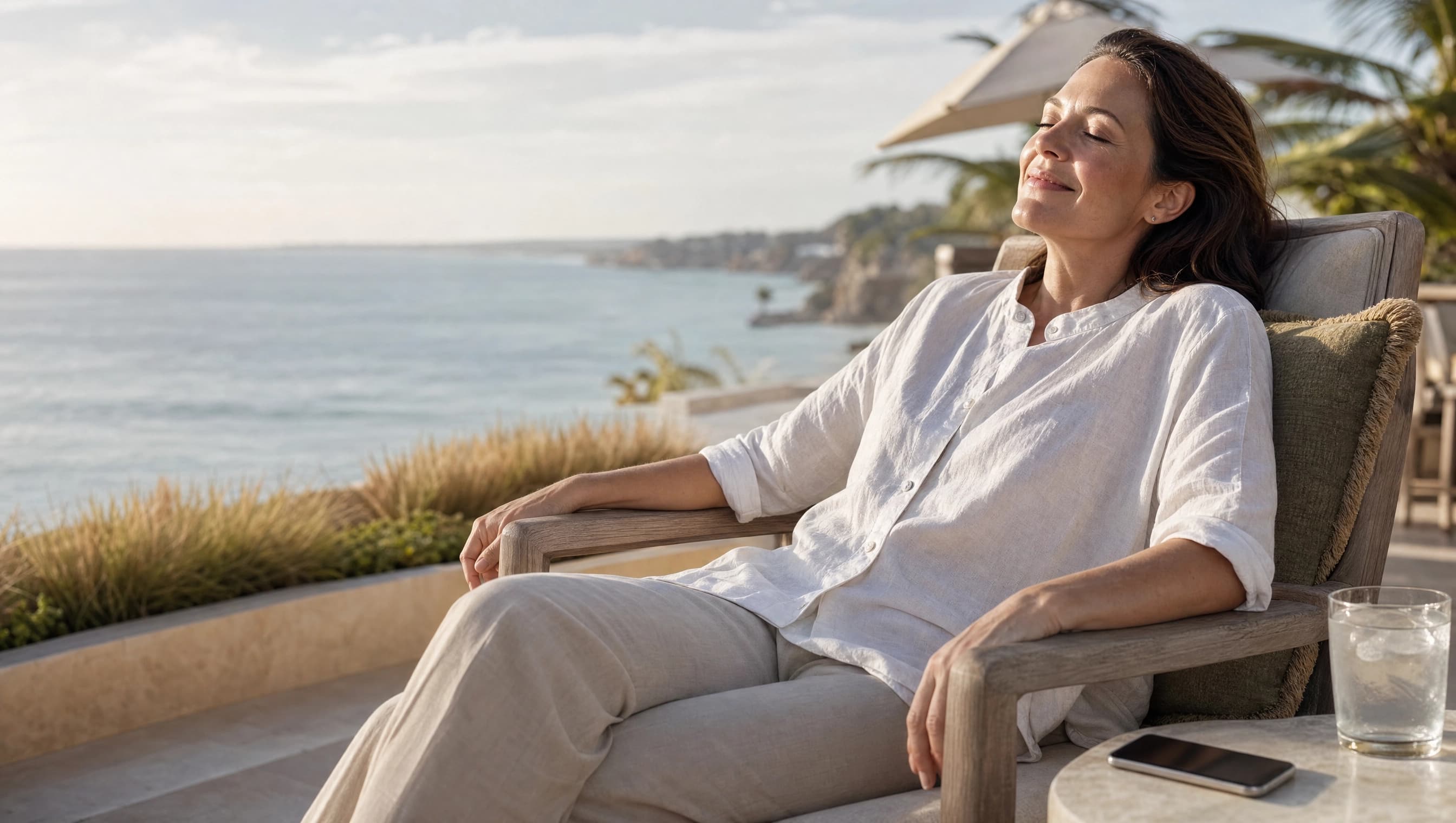 A relaxed professional woman with her eyes closed on an ocean-view terrace, phone resting face-down on the table beside her